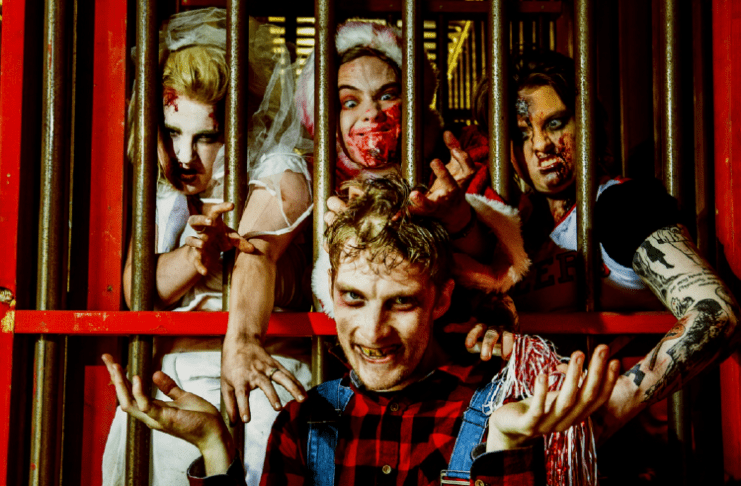 This is a photo of four scare actors in full makeup and costume. Three of them are trapped behind bars reaching towards the fourth.