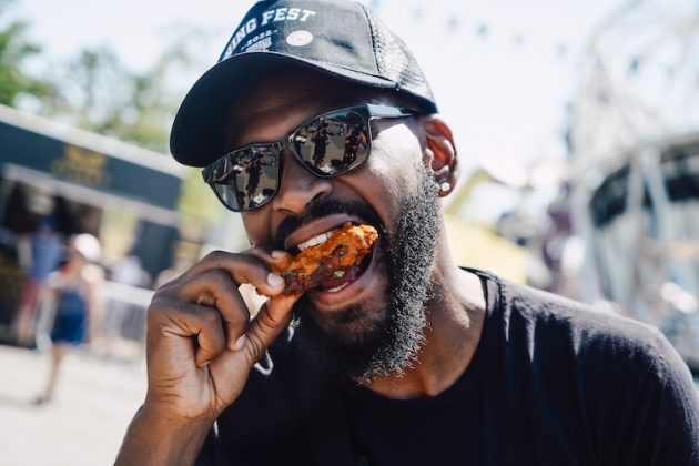 A man enjoying a piece of wing wearing sunglasses and a cap.