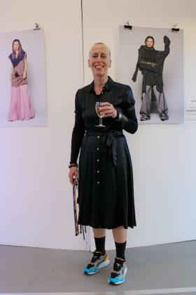 Fiona Hawthorne, the organiser of the Fashion Show in Derby and senior lecturer on the BA Fashion Studies standing in front of photos of students' outfits with glass of wine.