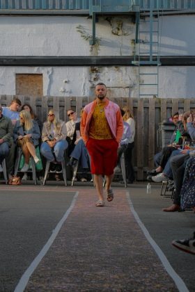 A male model walking down the runway wearing red shorts and yellow shirt with pink jacket.