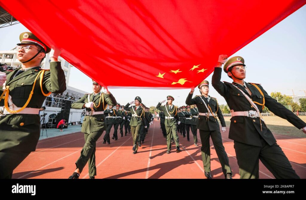 HUAI'AN, CHINA - Freshmen from Huaiyin Normal University perform a military training demonstration in Huai 'an, Jiangsu province. They are wearing green military-style uniforms and four of them are holding a red Chinese flag with five gold stars. The freshmen are marching under the flag, which they hold with one hand each.