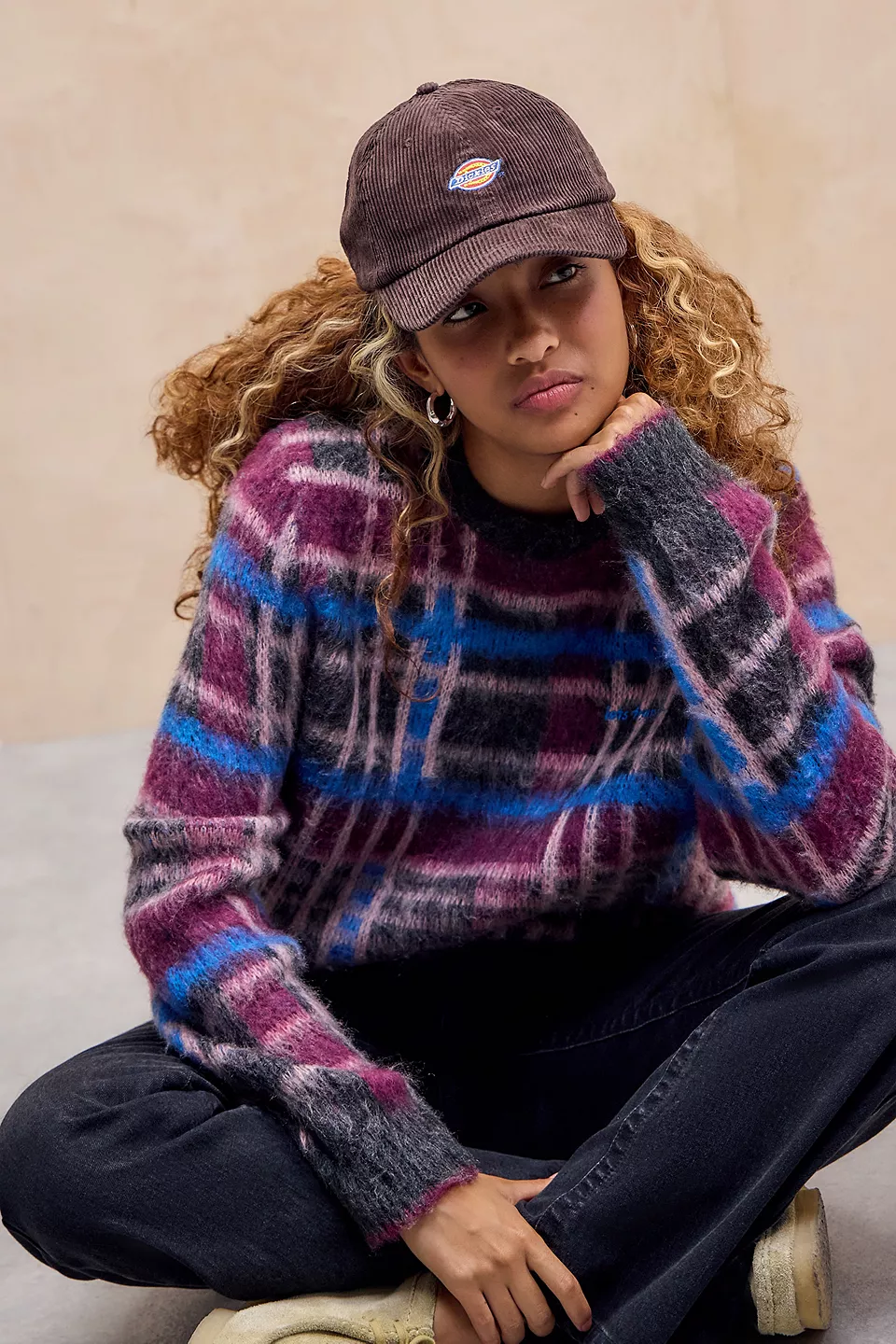 a person wearing a cozy plaid sweater in pink, purple and blue tones. paired with dark jeans and a brown cap