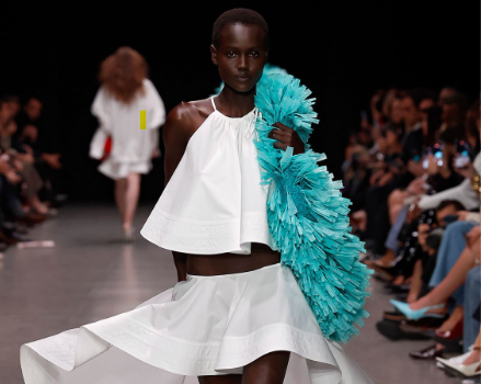 Model wearing Stella McCartney runway outfit featuring a crisp white top with bold blue ruffles, paired with a white skirt that is cut at the front yet flows into a longer draped back, modern yet elegant and sustainable