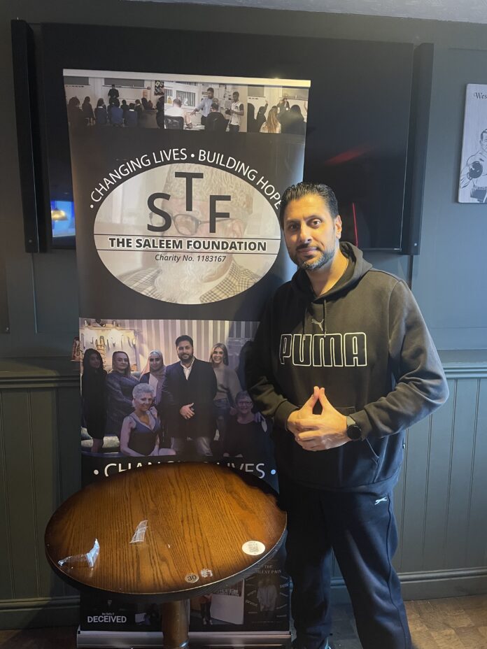 This image shows Shaz Saleem, a charity founder and Conservative councillor, standing in front of a banner that shows his charity - The Saleem Foundation.